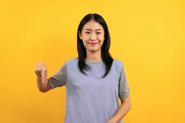 広告商品か何かを指差すために前に指を行うカジュアルな服の若いアジアの女性や幸福が黄色の背景に隔離されていると笑顔 — ストック写真