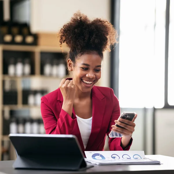 Takım elbiseli Afro-Amerikan iş kadını akıllı telefondan iyi haberler okuyor ve modern çalışma ortamında dijital tablet ve evrak işleriyle uğraşırken başarılı bir şekilde çalışmak için yumruğunu kaldırıyor..