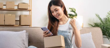 Genç girişimci akıllı telefonu online siparişleri kontrol etmek ve satış ürünlerinde başarılı bir şekilde kollarını kaldırırken aynı zamanda kanepede rahat bir şekilde oturuyor ve merkez ofisteki oturma odasında çalışıyor..