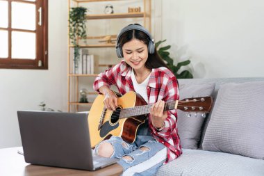 Genç kadın gitar çalma ve kulaklık takıp dizüstü bilgisayarla müzik aleti çalarken oturma odasındaki kanepede rahat bir şekilde oturmaya çalışıyor..