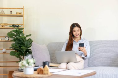 Serbest çalışan bir kadın akıllı telefondan iş verilerini okuyor ve dizüstü bilgisayarında bilgi yazarken aynı zamanda koltukta oturup not defteri üzerinde notlar alıyor ve ev ofisinin oturma odasında çalışıyor..