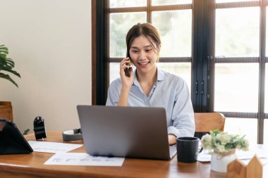 Kadın serbest meslek arkadaşıyla akıllı telefondan konuşuyor ve merkez ofisteki oturma odasındaki masada çalışırken laptopta bilgi yazıyor..