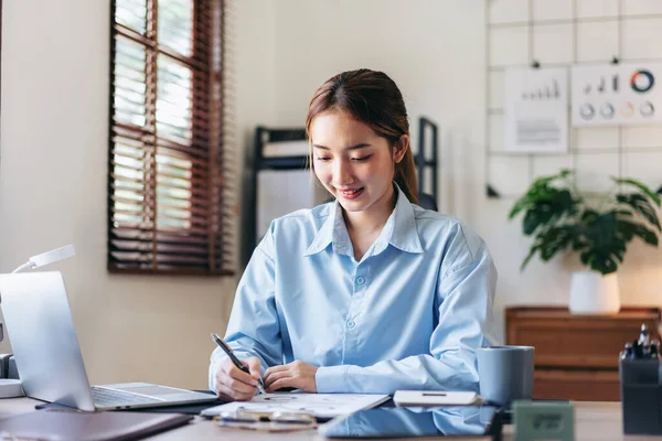 Businesswoman is reading information of new business project and taking notes in document while thoughtful about accounting of business and working on laptop at modern office.