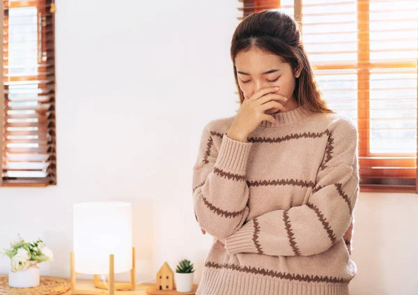 Woman in sweater covering nose with hand while feeling sick and headache with migraine during resting and standing in living room at home.