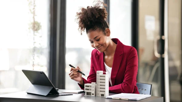 African american businesswoman in suit pointing condominium model to presenting and working on digital tablet with contract document about renting or buying and insurance housing in workspace modern.