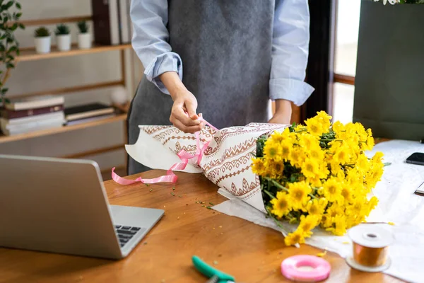 Female florist in apron using craft paper to wrapping yellow chrysanthemum and tie with pink ribbon on the table while creating and designing floral for arrangement flower bouquet in her flower shop.