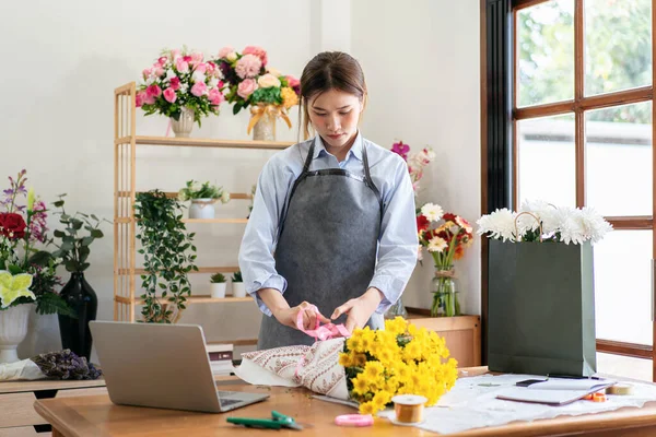 Female florist in apron using craft paper to wrapping yellow chrysanthemum and tie with pink ribbon on the table while creating and designing floral for arrangement flower bouquet in her flower shop.