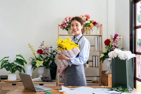 Female florist in apron holding yellow chrysanthemum bouquet wrapping with craft paper and tie with pink ribbon after creating and designing floral for arrangement flower bouquet in her flower shop.