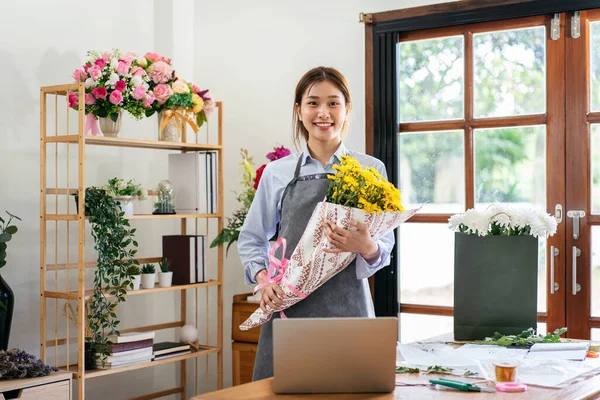 Female florist in apron holding yellow chrysanthemum bouquet wrapping with craft paper and tie with pink ribbon after creating and designing floral for arrangement flower bouquet in her flower shop.