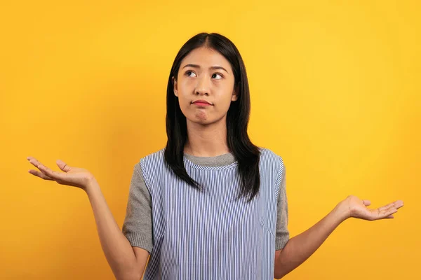 Young asian woman in casual clothes raising to open hands and confused face while gesturing doubting and uncertain isolated over yellow background.