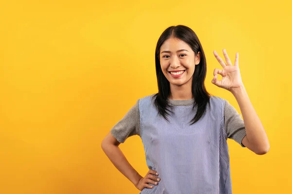 若いですアジアの女性でカジュアル服で手を腰に置くと笑顔と作りますOkジェスチャーで確認するために良い製品のために単離された黄色の背景 — ストック写真
