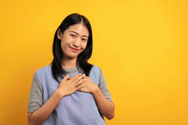 若いですアジアの女性でカジュアル服笑顔と胸の上に手を取ります幸せと感謝のジェスチャーを感じ黄色の背景に隔離 — ストック写真