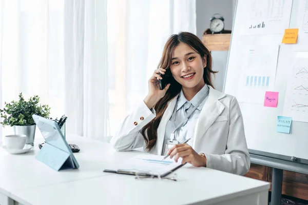 Takım elbiseli bir iş kadını, ortaklarıyla konuşmak için akıllı telefonu kullanır ve iş belgelerini okurken evrak işleri ile ilgili notlar alır ve modern ofisteki teknoloji cihazının tabletiyle çalışır..
