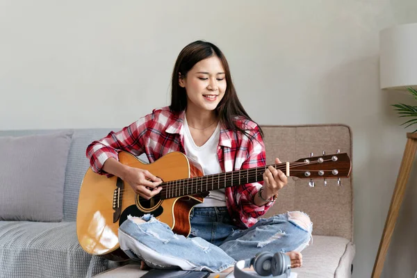 Young woman is practice basic guitar chords and playing guitar with pop songs while sitting on comfortable the couch in living room at home.