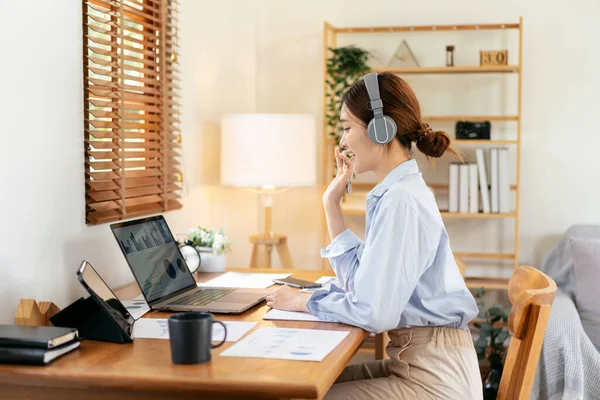 Female freelance is using laptop and wearing headphone to meeting video conference with colleague while working on the table in living room at home office.