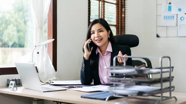 Young businesswoman in suit is talking with partner on smartphone while working about analysis of strategy and planning new business in office.