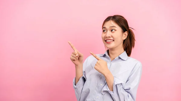若いアジアの女性は長い袖のシャツを着て ピンクの背景に隔離されたコピースペースに広告を指すために2本の額で手を使用して — ストック写真