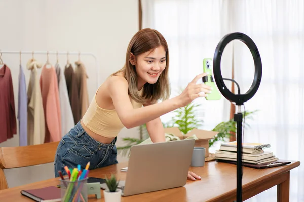 Woman designer entrepreneur preparing smartphone and device to showing clothes on live online social media streaming and packing product into cardboard box to shipping customer delivery at home.