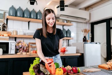 Elinde domates tutan bir kadın taze sebze ve organik ürünlerle evde modern mutfakta sağlıklı yemek pişirmek için sofrada meyveler hazırlarken keyif çatıyor..