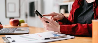 Genç iş kadını iş bilgilerini araştırmak için akıllı telefonu kullanıyor ve evde mutfaktaki masada dizüstü bilgisayarla çalışırken yeni iş projesini düşünüyor..