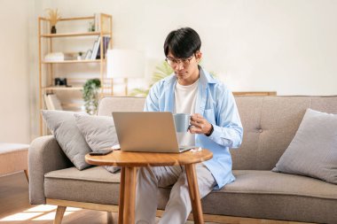 Gündelik iş adamı rahat koltukta oturup yeni iş için düşünceli bir şekilde sıcak çikolata içerken aynı zamanda iş projesiyle ilgili bilgi yazmak ve çalışmak için bilgisayarını kullanıyor..