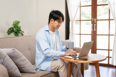 Gündelik iş adamı rahat koltukta oturup yeni iş için düşünceli bir şekilde sıcak çikolata içerken aynı zamanda iş projesiyle ilgili bilgi yazmak ve çalışmak için bilgisayarını kullanıyor..