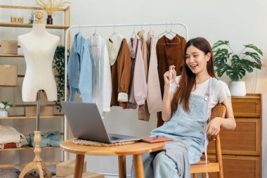 Kadın çevrimiçi satıcı, ölçü bandı tutarken ve müşteriyle sosyal medyada çevrimiçi kıyafet satmak için konuşurken yeni modalar tanıtmak için Vlog üzerinden canlı yayın yapmak için dizüstü bilgisayar kullanıyor.