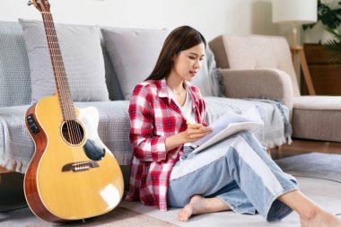 Genç kadın, evdeki oturma odasında yerde otururken gitar çaldıktan sonra şarkıyı ve şarkı sözlerini deftere yazıyor..