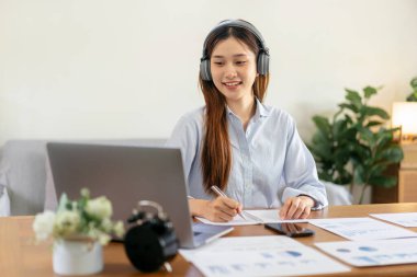 Kadın serbest yazar, müzik dinlemek için kulaklık takarken dizüstü bilgisayarda iş verilerini okuyor ve not defterine bilgi yazıyor ve merkez ofisteki masada çalışıyor..