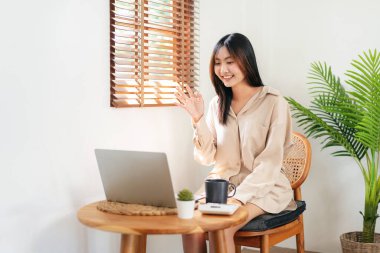 Kadın girişimci, video konferansında buluşmak ve evdeki oturma odasında dinlenmek için sandalyeye otururken meslektaşını karşılamak için dizüstü bilgisayar kullanıyor..