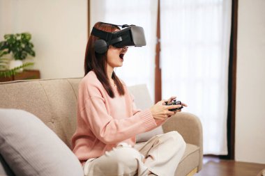 Evde oturma odasında dinlenirken elinde joystick ile sanal gerçeklikle video oyunu oynayan süveter ve sanal gözlük takan genç bir lezbiyen kadın..