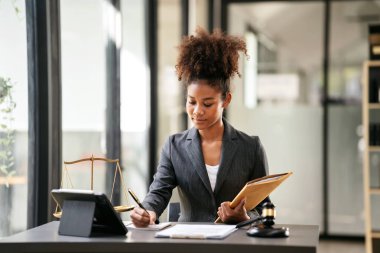 Takım elbiseli Afro-Amerikan avukat kadın elinde iş sözleşmesi zarfı tutarken ve not defterine bilgi yazarken aynı zamanda mahkeme masasında yargıç çekiciyle yasal ve adalet hakkında çalışıyor..