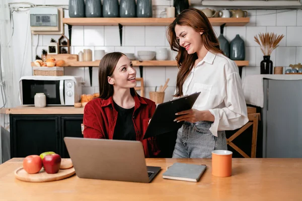 Young woman showing business information on clipboard to friend and reading data together to brainstorming about new project business while working with laptop on the table in kitchen at home.