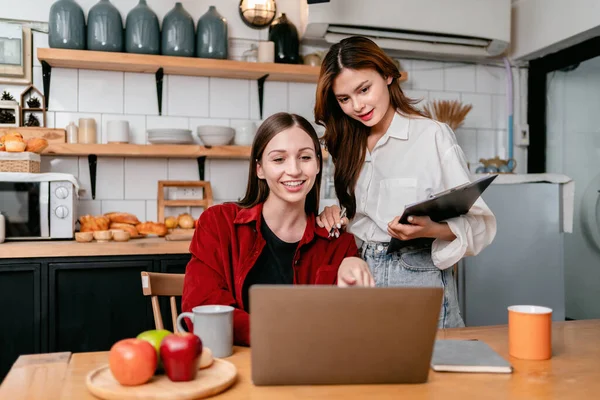 两名年轻妇女在家里厨房里一起工作时 用手提电脑上的数据和阅读商业信息来集思广益 — 图库照片