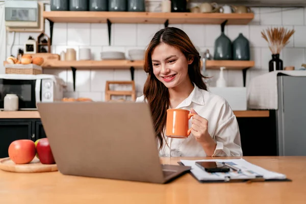 年轻的女商人一边坐着一边喝咖啡和打字 一边在笔记本电脑上搜索商业信息 同时思考着家里厨房里新的商业项目的策略 — 图库照片