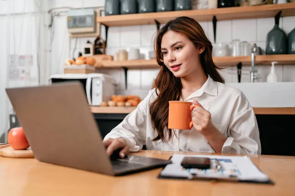 年轻的女商人一边坐着一边喝咖啡和打字 一边在笔记本电脑上搜索商业信息 同时思考着家里厨房里新的商业项目的策略 — 图库照片