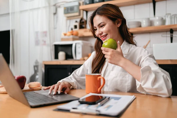 年轻的女商人一边吃着青苹果 一边在笔记本电脑上打字搜索商业信息 一边坐着工作 思考着家里厨房里新的商业项目的策略 — 图库照片