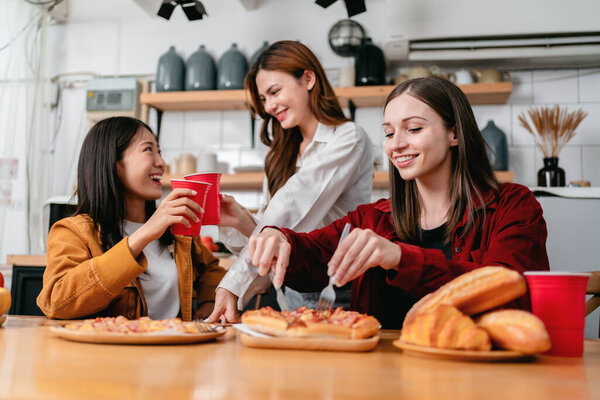 Group of friends clinking glass of beverage and cutting slice of italian pizza for dinner meal while having enjoying with party to celebration for weekend in the kitchen at home.