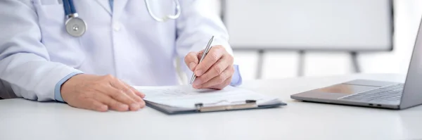 Hastanedeki dizüstü bilgisayarında hastalık bilgilerinin tedavisini ararken hasta hastalıklarına stetoskop teşhisi koyan ve panoya reçete yazan kıdemli erkek doktor..