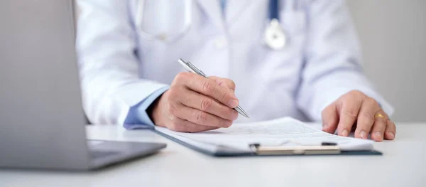stock image Senior male doctor with stethoscope diagnosis of patient disease and writing prescription on clipboard while working to searching treatment of disease information on laptop in hospital.