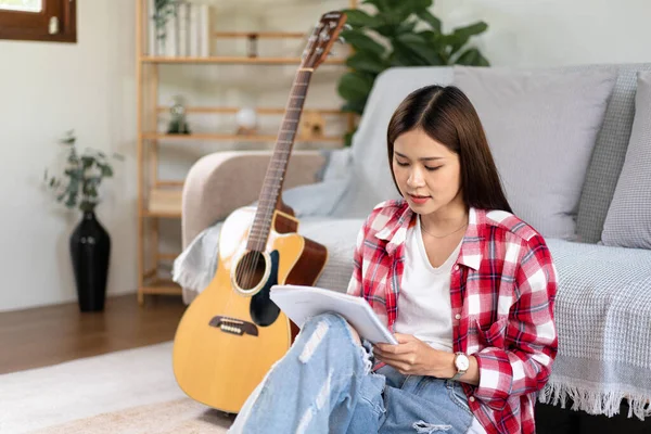 Young woman is compose the song and writing lyrics on notebook after playing guitar while sitting on the floor in living room at home.