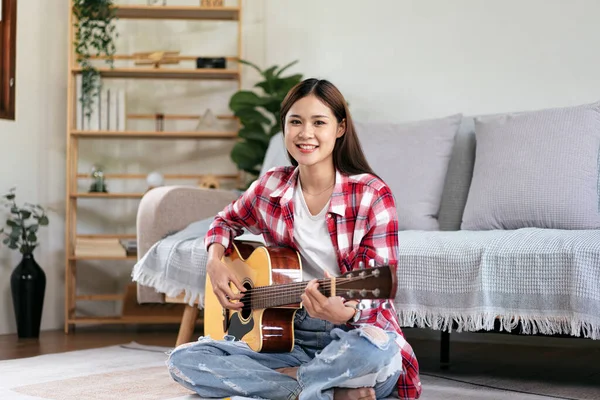 Young woman is playing guitar and practice to singing the song after compose a song while sitting on the floor in living room at home.