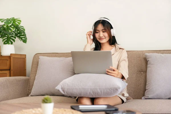 女企业家坐在舒适的大沙发上 把笔记本电脑放在枕头上 头戴耳机听音乐 看电影 在家里客厅里休息 — 图库照片