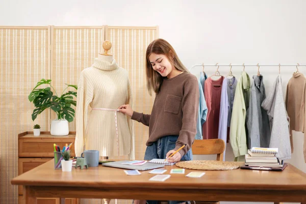 Woman designer entrepreneur using measure tape to measuring size sweater in mannequin and writing data in notebook while working to sketching design new collection for winter in home workshop.
