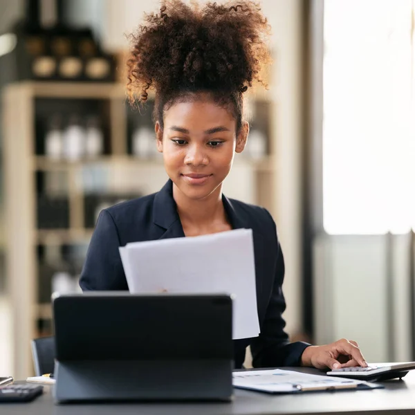 アフリカ系アメリカ人のビジネスウーマンは 現代のワークスペースで仕事をしながら 書類事務におけるビジネスの会計情報を読み 計算機で新しいビジネスの会計を計算する — ストック写真