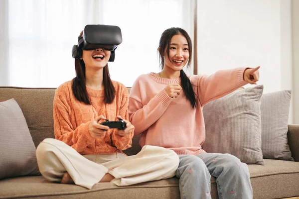 Young woman lesbian couple wearing sweater and VR goggles while holding joystick to playing video game with experience virtual reality and spending time together in living room at home.