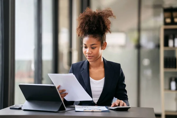 アフリカ系アメリカ人のビジネスウーマンは 現代のワークスペースで仕事をしながら 書類事務におけるビジネスの会計情報を読み 計算機で新しいビジネスの会計を計算する — ストック写真