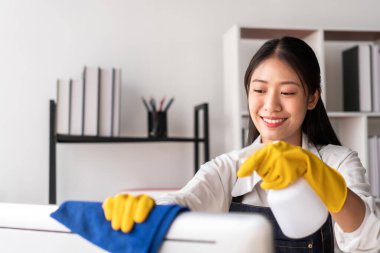 Önlüklü ev kadını, bilgisayara sprey sıkmak için eldiven giyiyor ve evde çalışırken temizlik yaparken temizlik yapmak için mikrofiber kumaş kullanıyor..