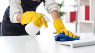 Önlüklü ev hanımı, bilgisayara sprey sıkmak için eldiven giyiyor ve evde çalışırken masayı temizlemek ve mobilyaları temizlemek için mikrofiber kumaş kullanıyor..