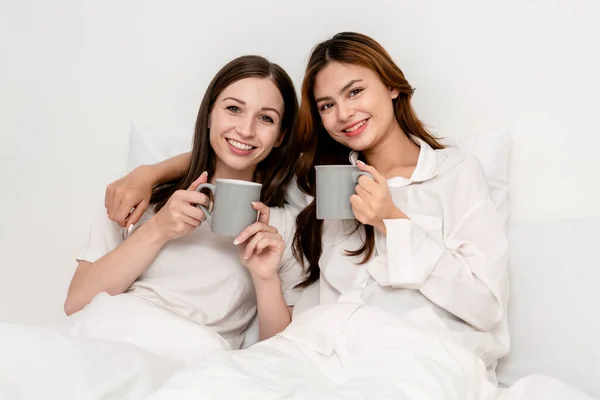 stock image Young asian couple woman drinking chocolate cup together while embracing and lying under blanket and leaning on comfortable pillows on the bed in bedroom at home.